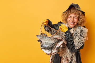 Horizontal shot of female angler returns from fishing trip caught different types of fish smiles toothily wears leather hat raincoat and rubber gloves isolated over yellow background blank space aside