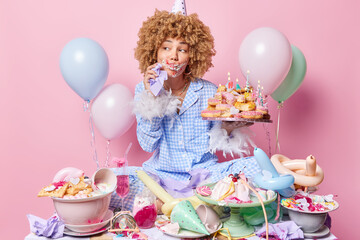 Obraz na płótnie Canvas Thoughtful curly haired young woman smeared with cream after eating cake wears cone hat and pajama holds tray with pile of doughnuts and burning candles poses near festive table full of desserts