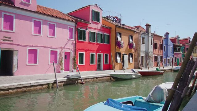Colorful semi-detached houses stand on bank of water channel with boats under bright sunlight and cloudless sky in Burano