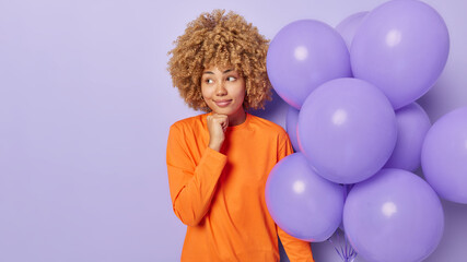Horizontal shot of thoughtful woman dressed in orange jumper keeps hand under chin concentrated aside holds bunch of inflated balloons thinks how to celebrate birthday isolated over purple background