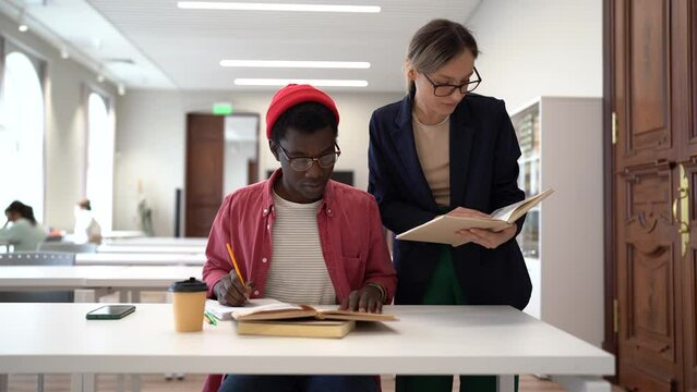 Two multiracial students studying together in library. Young diverse people sit at desk with books, working on collaborative research project. Teamwork and cooperative learning in higher education