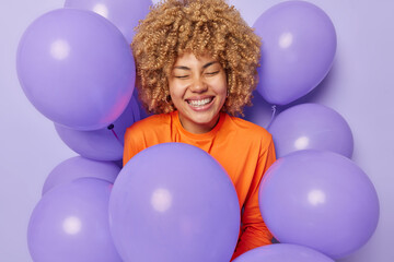 Fototapeta na wymiar Horizontal shot of cheerful woman poses around inflated balloons smiles broadly stands with closed eyes gets congratulations on birthday stands against purple studio background. Special occasion