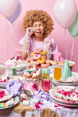 Unhappy doleful curly woman with unhappy expression wipes tears wears dress and silk gloves stands near messy table full of desserts surrounded by inflated balloons celebrates birthday alone