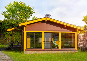 House with panoramic windows. One-story cottage with lawn. Wooden house in summer weather. Residential cottage with bicycle on terrace. Hedge behind country house. Scandinavian style cottage