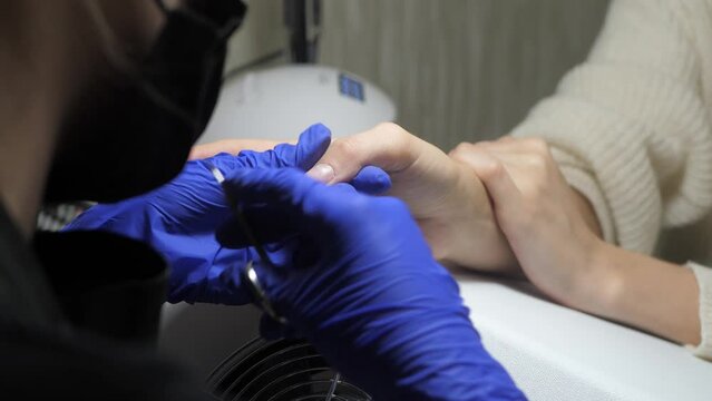Nail master in blue rubber gloves preparating of nails for manicure. Beautician cutting the cuticle by professional metal instrument in beauty salon. Cuticle removal by scissors