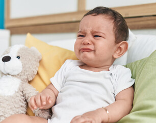 Adorable hispanic baby sitting on bed crying at bedroom