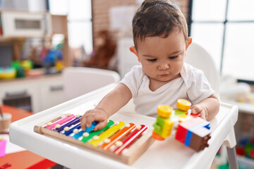 Adorable hispanic baby sitting on highchair playing xylophone holding car at kindergarten
