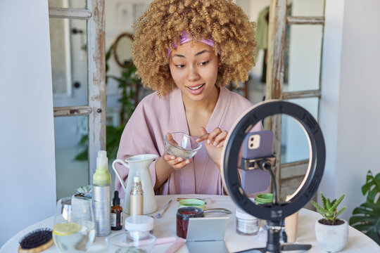 Indoor shot of curly haired adult woman dressed in bathrobe records video blog tells about cosmetic products for your beauty poses against blurred home interior. Blogging and cosmetology concept