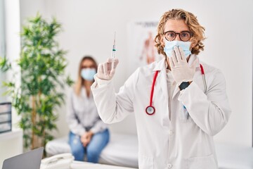 Blond man wearing doctor uniform and medical mask holding syringe covering mouth with hand, shocked and afraid for mistake. surprised expression