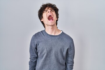 Young man standing over isolated background angry and mad screaming frustrated and furious, shouting with anger. rage and aggressive concept.