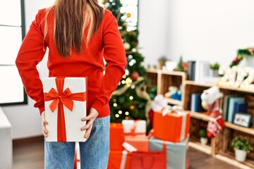 Young caucasian woman holding gift standing by christmas tree at home