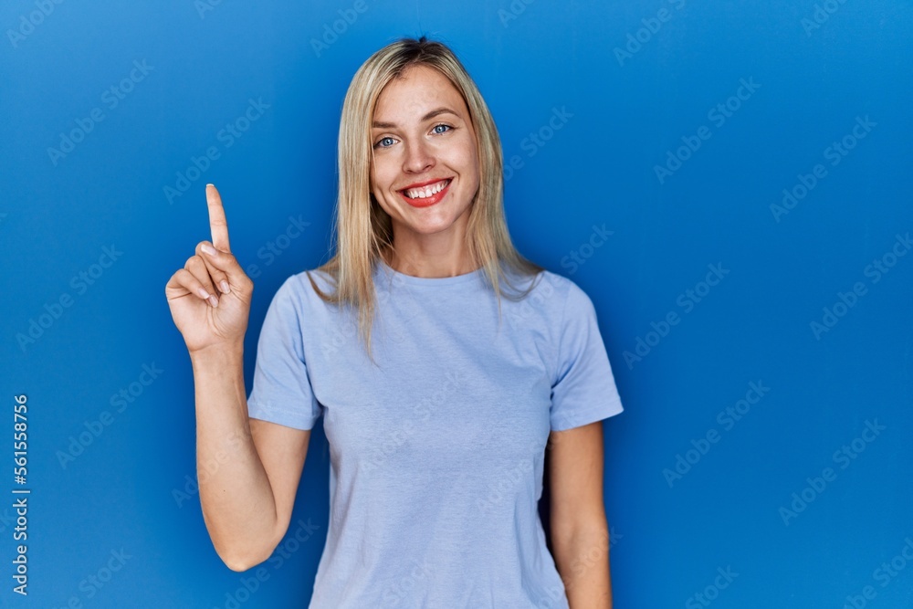 Poster beautiful blonde woman wearing casual t shirt over blue background showing and pointing up with fing