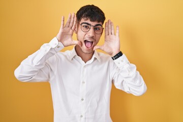 Young hispanic man standing over yellow background smiling cheerful playing peek a boo with hands...