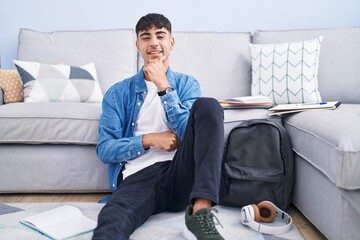 Young hispanic man sitting on the floor studying for university with hand on chin thinking about question, pensive expression. smiling and thoughtful face. doubt concept.