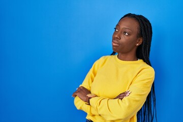 Beautiful black woman standing over blue background looking to the side with arms crossed convinced and confident