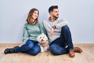 Young hispanic couple sitting on the floor with dog looking away to side with smile on face, natural expression. laughing confident.