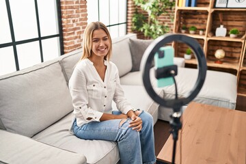 Young blonde woman recording vlog tutorial with smartphone at home with a happy and cool smile on...