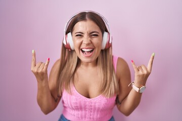 Young blonde woman listening to music using headphones shouting with crazy expression doing rock symbol with hands up. music star. heavy music concept.