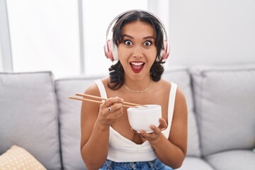 Young hispanic woman eating asian food using chopsticks afraid and shocked with surprise and amazed expression, fear and excited face.