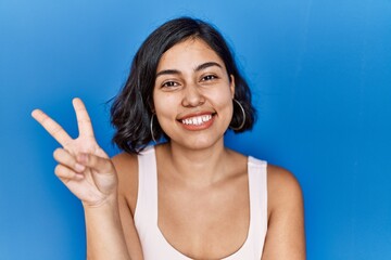 Fototapeta premium Young hispanic woman standing over blue background smiling with happy face winking at the camera doing victory sign. number two.