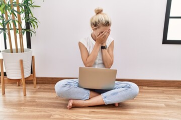 Young blonde woman using computer laptop sitting on the floor at the living room with sad...