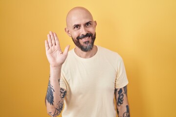 Hispanic man with tattoos standing over yellow background waiving saying hello happy and smiling, friendly welcome gesture