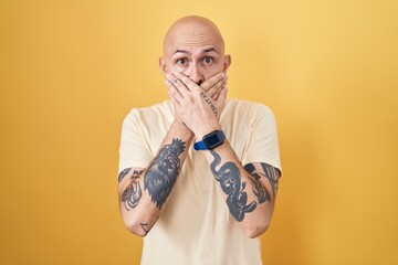 Hispanic man with tattoos standing over yellow background shocked covering mouth with hands for...
