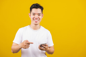 Asian handsome young man smiling positive holding smartphone blank screen and pointing finger to mobile phone, studio shot isolated on yellow background, making successful expression gesture concept