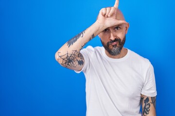 Hispanic man with tattoos standing over blue background making fun of people with fingers on...