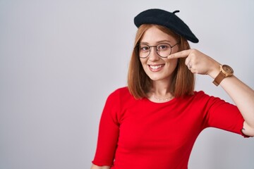 Young redhead woman standing wearing glasses and beret pointing with hand finger to face and nose, smiling cheerful. beauty concept