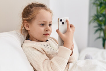 Adorable blonde girl using smartphone sitting on bed at bedroom