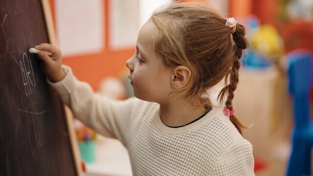 Adorable blonde girl preschool student drawing on blackboard at kindergarten