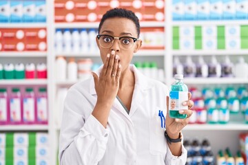 African american woman working at pharmacy drugstore holding syrup covering mouth with hand, shocked and afraid for mistake. surprised expression