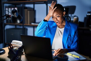 Beautiful african american woman working at the office at night surprised with hand on head for mistake, remember error. forgot, bad memory concept.