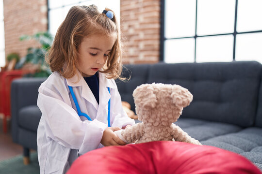 Adorable Hispanic Girl Wearing Doctor Uniform Putting Band Aid On Teddy Bear Arm At Home