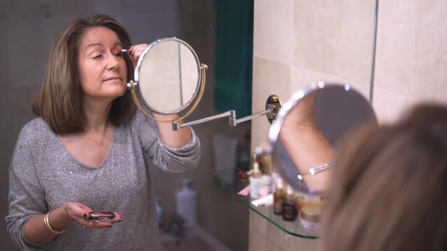 Senior White Woman Putting On Makeup In Front Of The Mirror In Her Bathroom.