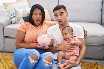 Young hispanic couple with baby holding piggy bank in shock face, looking skeptical and sarcastic,...