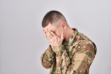 Young man wearing camouflage army uniform with sad expression covering face with hands while...