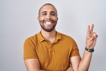 Hispanic man with beard standing over white background smiling with happy face winking at the camera doing victory sign with fingers. number two.