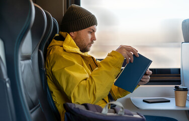 Bearded man in winter yellow jacket and hat reading book while traveling by train sitting inside...