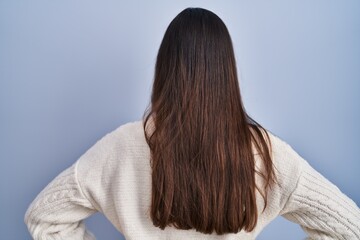 Young brunette woman standing over blue background standing backwards looking away with arms on body