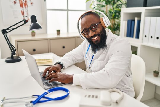 Young African American Man Wearing Doctor Uniform Using Laptop And Headphones Working At Clinic