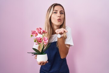 Young blonde woman wearing gardener apron holding plant looking at the camera blowing a kiss with hand on air being lovely and sexy. love expression.