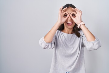 Young hispanic woman standing over white background doing ok gesture like binoculars sticking tongue out, eyes looking through fingers. crazy expression.