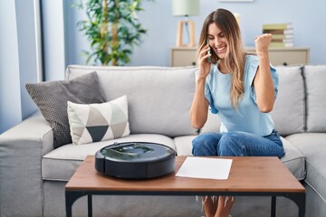 Young hispanic woman sitting at home by vacuum robot speaking on the phone screaming proud,...