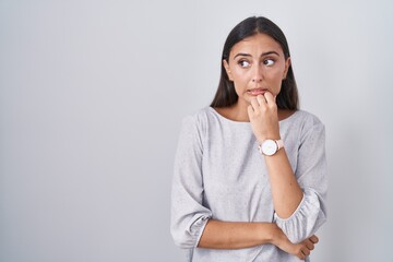 Young hispanic woman standing over white background looking stressed and nervous with hands on mouth biting nails. anxiety problem.