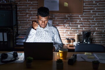 Fototapeta premium Young hispanic man working at the office at night angry and mad raising fist frustrated and furious while shouting with anger. rage and aggressive concept.