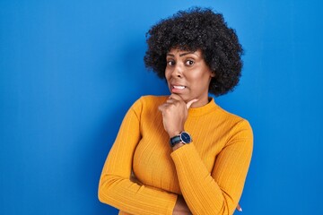 Fototapeta na wymiar Black woman with curly hair standing over blue background thinking worried about a question, concerned and nervous with hand on chin