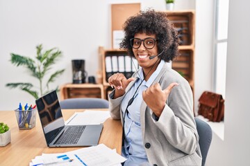 Black woman with curly hair wearing call center agent headset at the office pointing to the back behind with hand and thumbs up, smiling confident