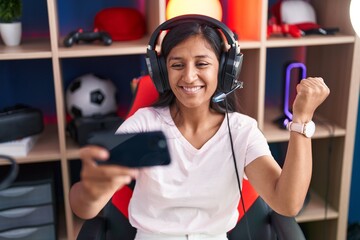 Young hispanic woman playing video games with smartphone screaming proud, celebrating victory and...